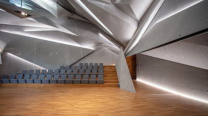 Ein futuristisch wirkender Raum mit ineinander verschachtelten Elementen mit hellgrauen Stuhlreihen und Parkettboden