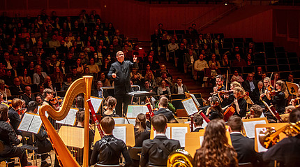 Ein Herr im schwarzen Anzug steht vor einem Orchester und dirigiert.
