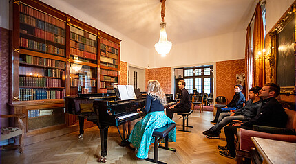 Eine Dame sitzt in einem historischen Raum und spielt am Flügel. Vier Personen sitzen um sie herum und hören zu.