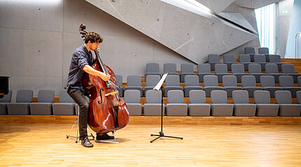 Ein junger Mann mit einem Kontrabass spielt mit seinem Instrument in einem Raum mit Gesteinselementen. Vor ihm steht ein Notenständer. Hinter ihm sind graue gepolsterte Sitzreihen.