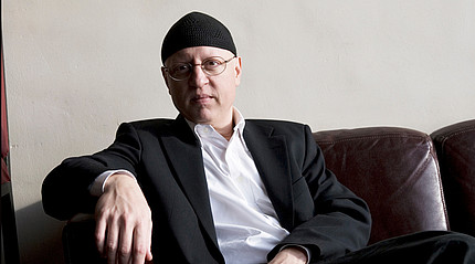 Ein Mann mit einer Mütze sitzt auf einem Sofa und blickt in die Kamera