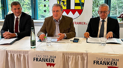 Drei Männern mit Anzügen sitzen an einem Tisch, im Vordergrund ist ein Logo zum Tag der Franken zu sehen