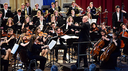 Ein Orchester mit jungen Leuten sitzt auf einer Bühne, ein Dirigent im dunklen Anzug steht im Vordergrund und hebt die Arme 