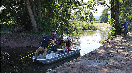 Auf einem Fluss liegt ein Boot, das von einer Frau am Motor gesteuert wird. Auf dem Boot sind zwei Männer mit Käschern und Käppies auf dem Kopf.