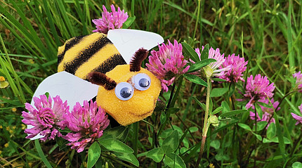 Eine gebastelte Biene sitzt im Gras zwischen lila Blüten