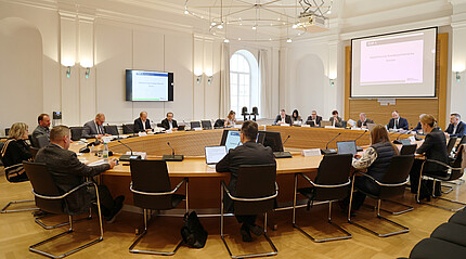 Sitzung im Großen Sitzungssaal des Bezirkstags In einem großen Saal sitzen an einem runden Konferenztisch Menschen. Vor sich haben Sie Unterlagen und Mikrofone auf dem Tisch.