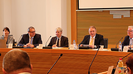 Vier Männer und eine Frau sitzen an einem Holztisch in einem Sitzungssaal
