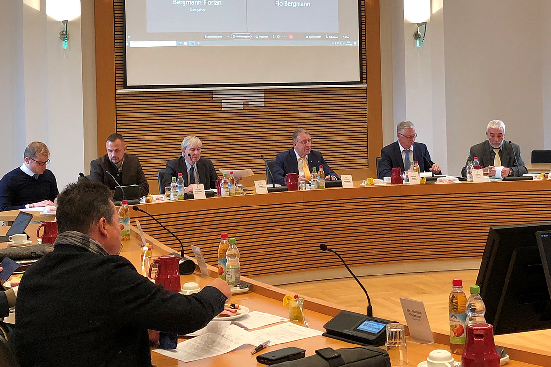 An einem halbrunden Sitzungstisch sitzen mehrere Männer