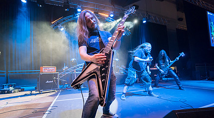 Ein Mann mit langen Haaren spielt auf einer großen Bühne im blauen Scheinwerferlicht auf einer E-Gitarre