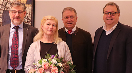 Drei Männer im Anzug und eine Frau mit Blumenstrauß stehen nebeneinander und blicken in die Kamera