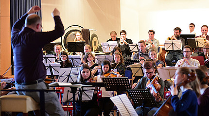Junge Musikerinnen und Musiker sitzen in einem Raum und proben. Im Vordergrund steht ein Dirigent.