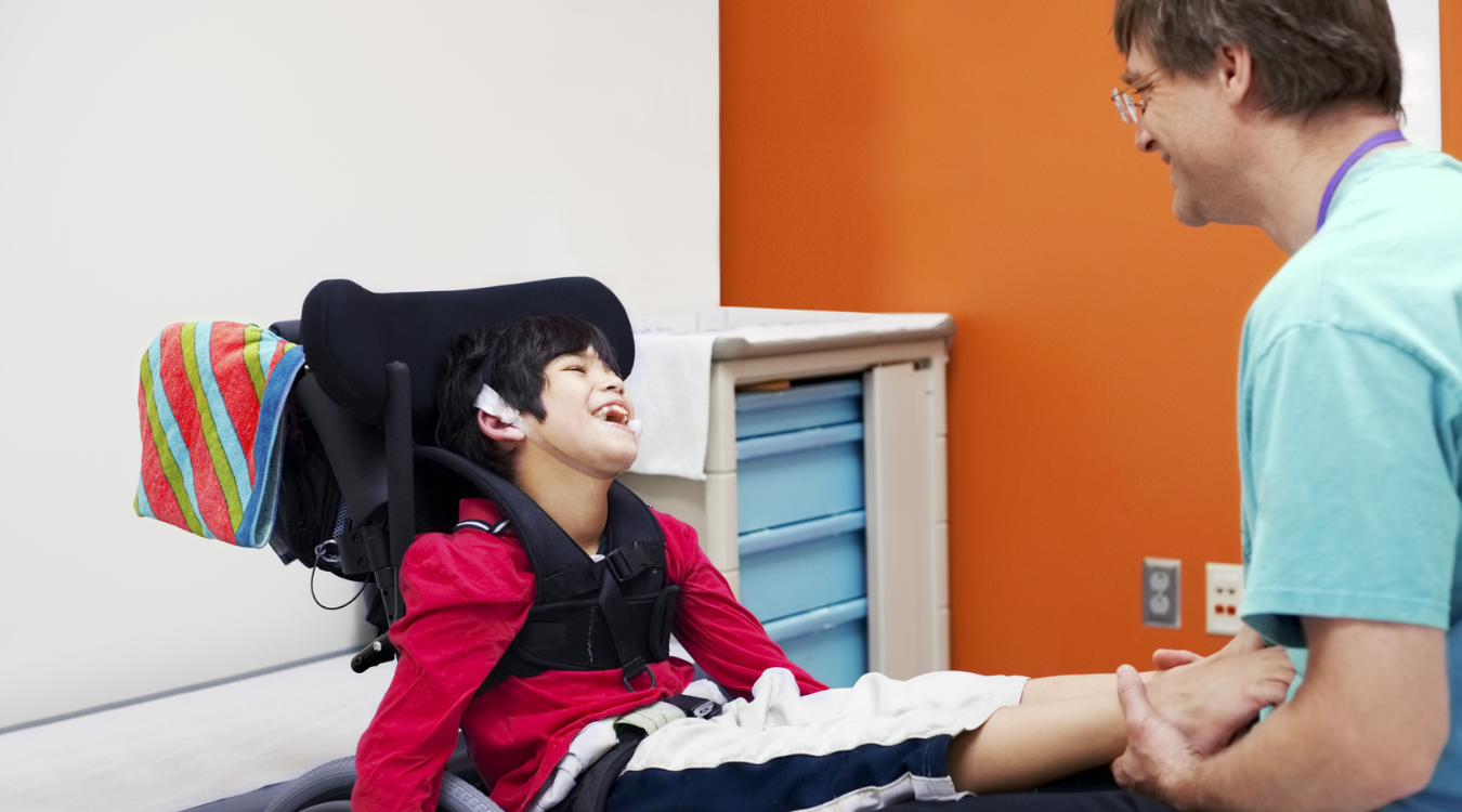Zu sehen ist ein behindertes Kind in einem Rollstuhl und ein Pfleger.