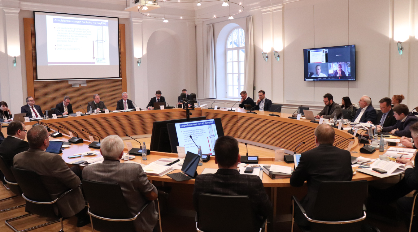 An einem großen runden Holztisch sitzen viele Menschen herum. Sie tragen Anzüge. Vor ihnen stehen Mikrofone. Auf mehreren Bildschirmen werden Zahlen und Diagramme gezeigt.