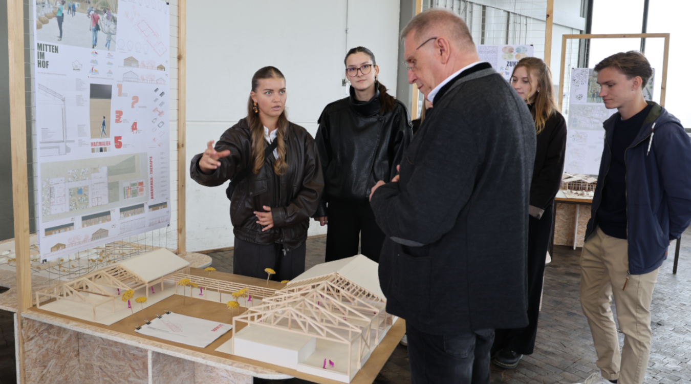 Eine junge Frau redet und gestikuliert vor einer Wand, an der Entwürfe hängen. Auf einem Tisch davor ist ein Holzmodell aufgebaut, das ein älterer Herr in einem Trachtenjancker betrachtet. Im Hintergrund stehen weitere junge Leute.