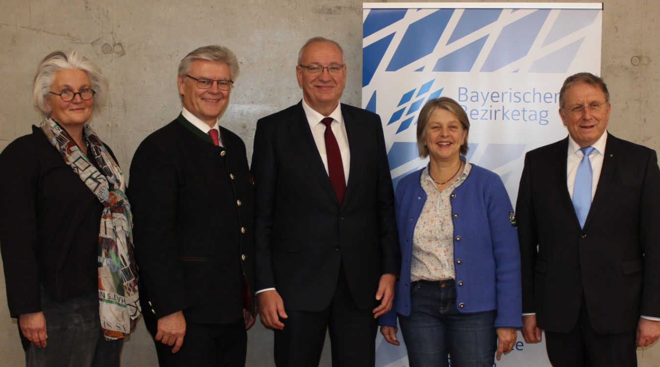 Drei Männer in dunklen Anzügen und zwei Frauen stehen in einer Reihe vor einer Wand.
