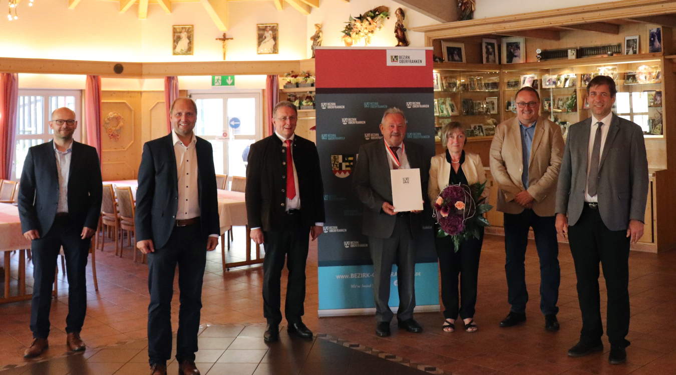 Bezirksmedaille in Silber für Franz Schmidtlein Sechs Männer und eine Frau stehen in einem Saal. Ein Mann hält ein Papier mit dem Bezirkswappen in den Händen und eine Frau einen Blumenstrauß.