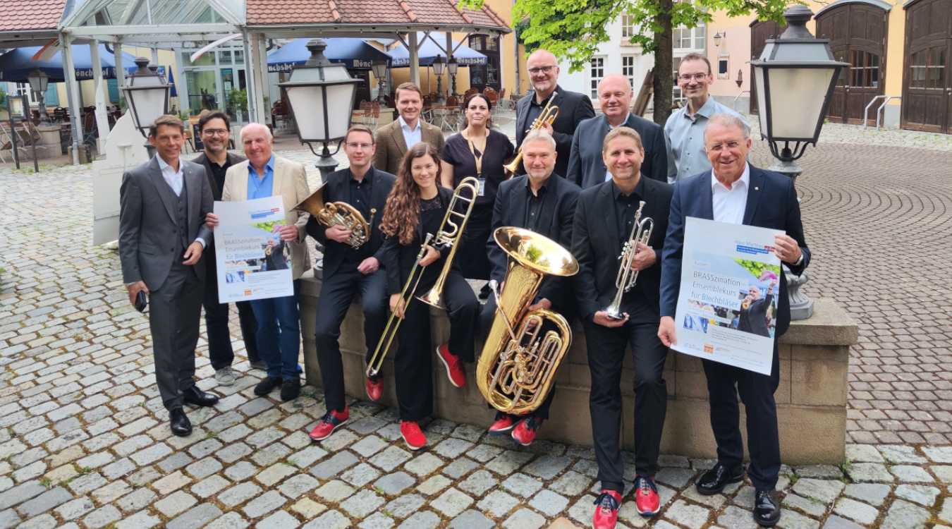 Eine Gruppe Menschen, einige mit einem Blechblasinstrument in der Hand, stehen in einem gepflasterten Innenhof.
