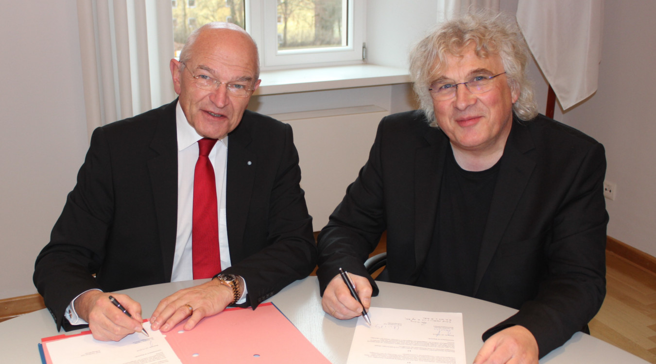 Bezirkstagspräsident Dr. Günther Denzler und der neue künstlerische Leiter von Haus Marteau, Prof. Christoph Adt, sitzen an einem runden Tisch und lächeln in die Kamera. Vor den Beiden liegen Mappen mit den Ausfertigungen des Vertrages. 