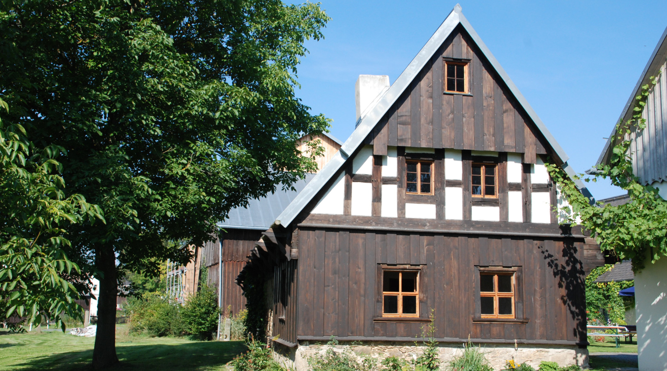 Denkmalpreis 2023 Ein saniertes kleines Haus mit Fachwerk und einer Holzfassade umgeben von einem Garten mit großen Bäumen