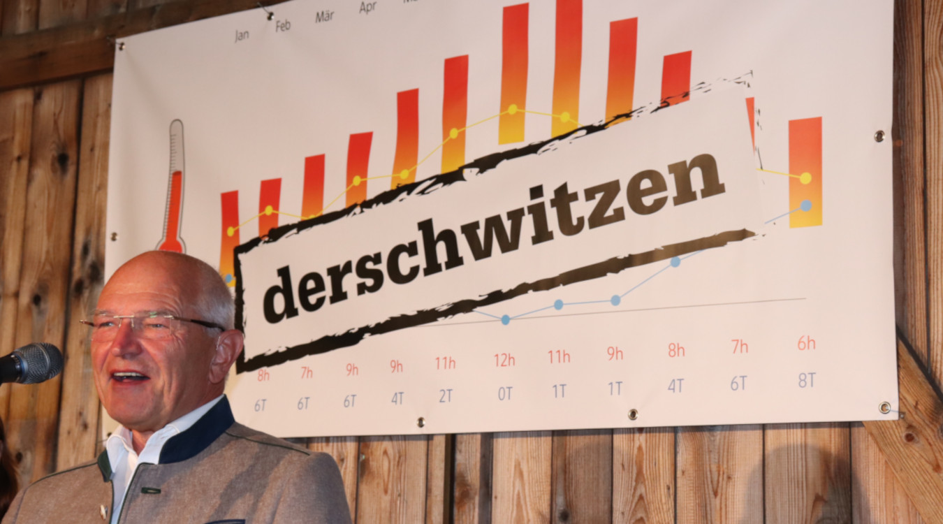 Das Bild zeigt Bezirkstagspräsident Dr. Günther Denzler vor einem Mikrofon. Hinter ihm hängt ein Plakat auf dem das Wort derschwitzen steht.