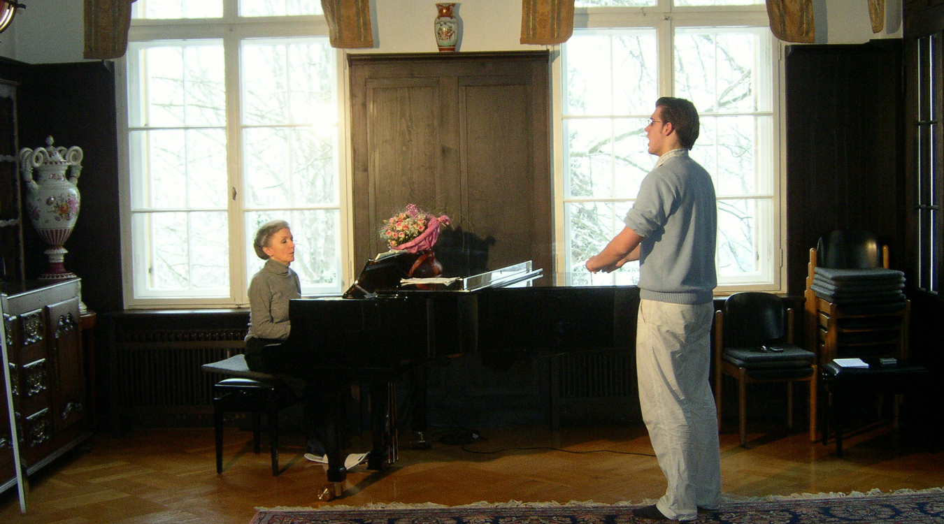 Eine ältere Dame sitzt am Klavier und leitet einen Sänger mittleren Alters an.