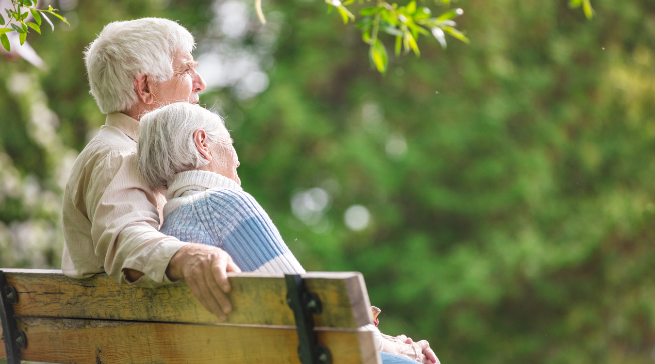 Ein alter Mann und eine Frau sitzen gemeinsam auf einer Bank. Die Frau in einem hellblauen Pullover legt ihren Kopf an die Schulter des Mannes.