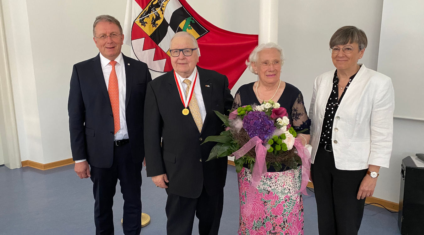 Ehrenmedaille in Gold für einen besonderen Repräsentanten Oberfrankens Zwei Männer und zwei Frauen stehen nebeneinander. Ein älterer Herr trägt eine goldene Medaille um den Hals. Eine ältere Frau hält einen Blumenstrauß in den Händen. Hinter den Personen ist die Fahne des Bezirks Oberfranken zu sehen.