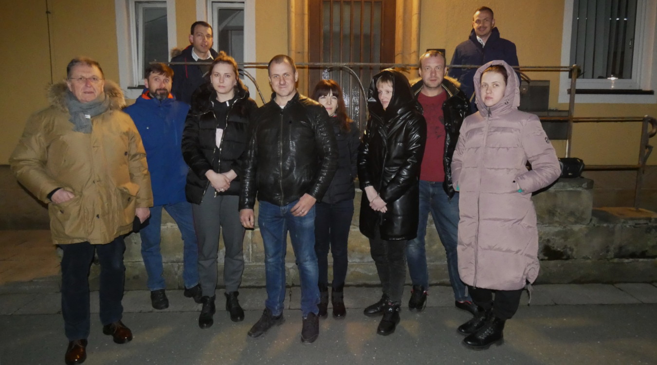 Vier Frauen und sechs Männer tragen dicke Jacken und stehen vor einem Haus in der Dunkelheit.