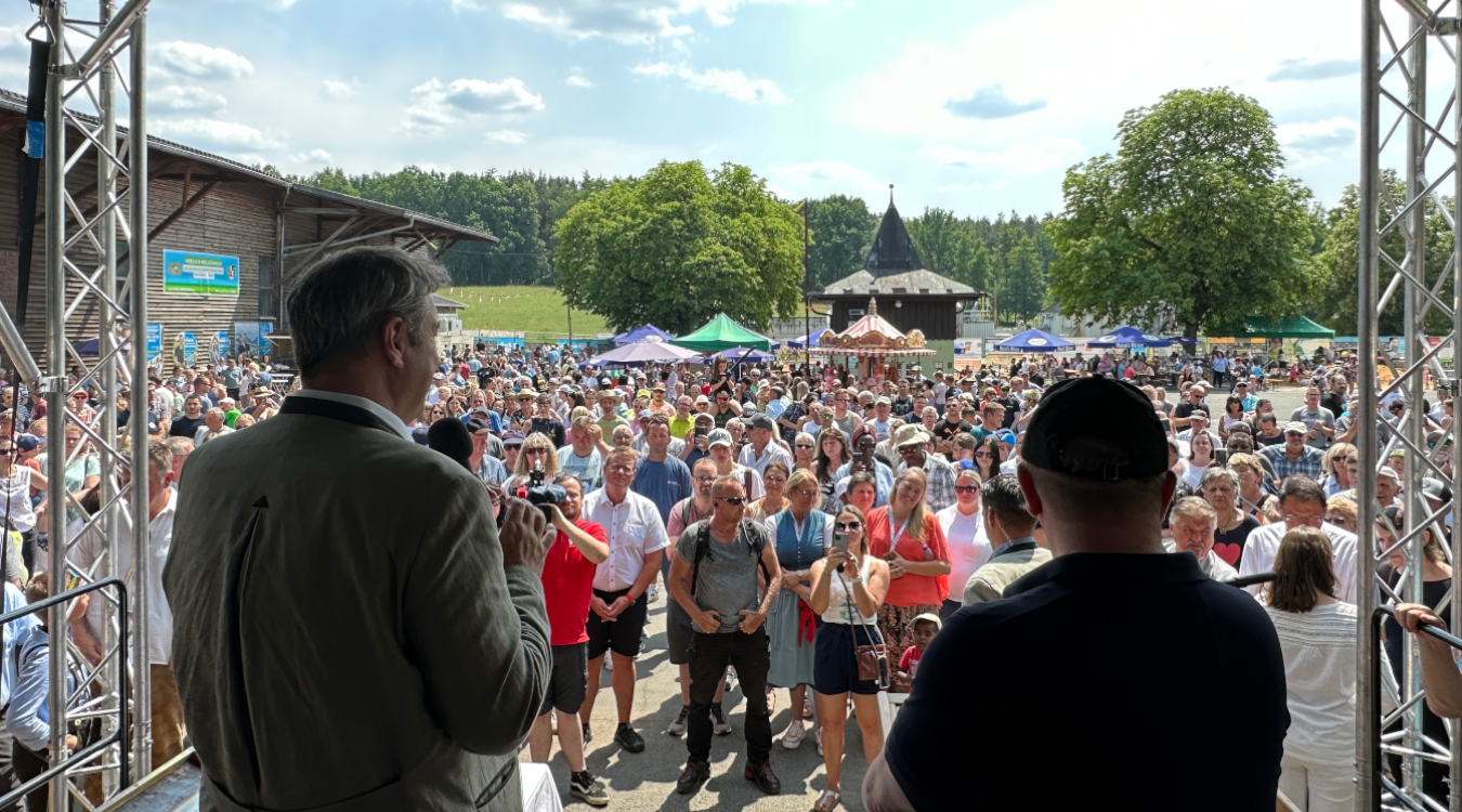Zwei Männer stehen auf einer Bühne, vor der Bühne stehen sehr viele Menschen auf einem Platz im Sonnenschein