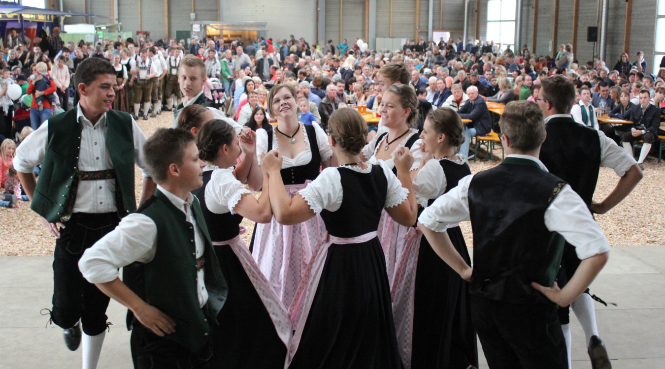 Die Landjugend Bad Berneck zeigt in der vollbesetzten Bodenhalle einen traditionellen Volkstanz. Die jungen Pärchen sind in einer schönen Tracht gekleidet. Im Hintergrund sind die Biertischgarnituren zu sehen, die allesamt mit Besuchern besetzt sind.