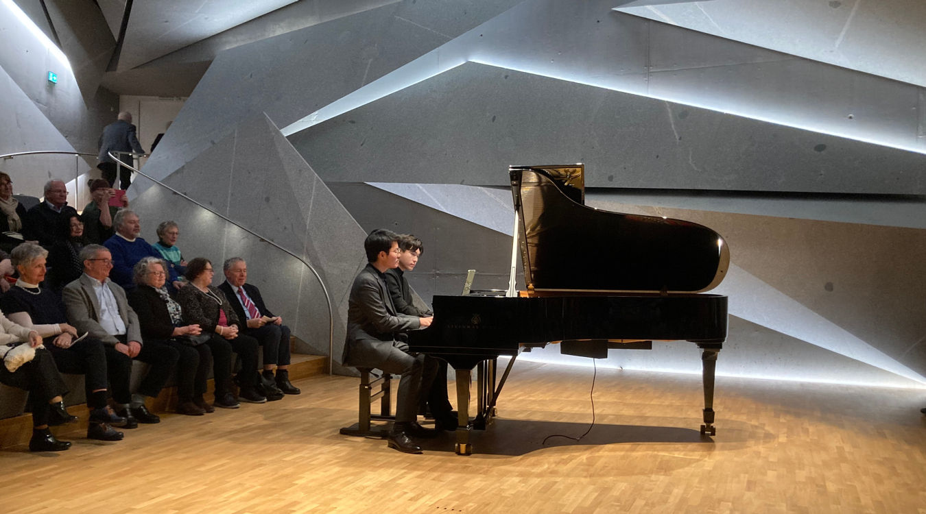 Junhee Kim und Honggi Kim In einem modern gestalteten Saal mit Gesteinsformen sitzen zwei junge Männer an einem Flügel und spielen. Auf Rängen hinter ihnen sitzen Zuschauer.