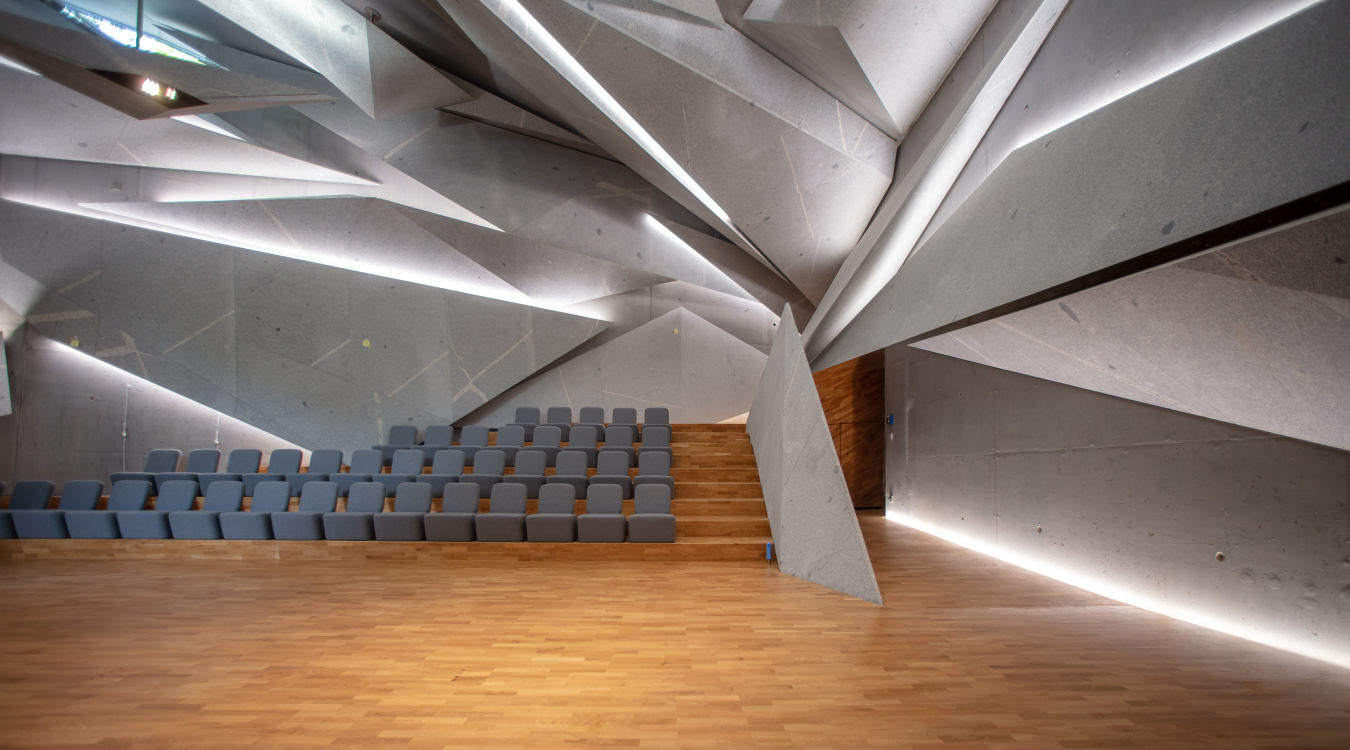 Konzertsaal Haus Marteau Ein futuristisch wirkender Raum mit ineinander verschachtelten Elementen mit hellgrauen Stuhlreihen und Parkettboden