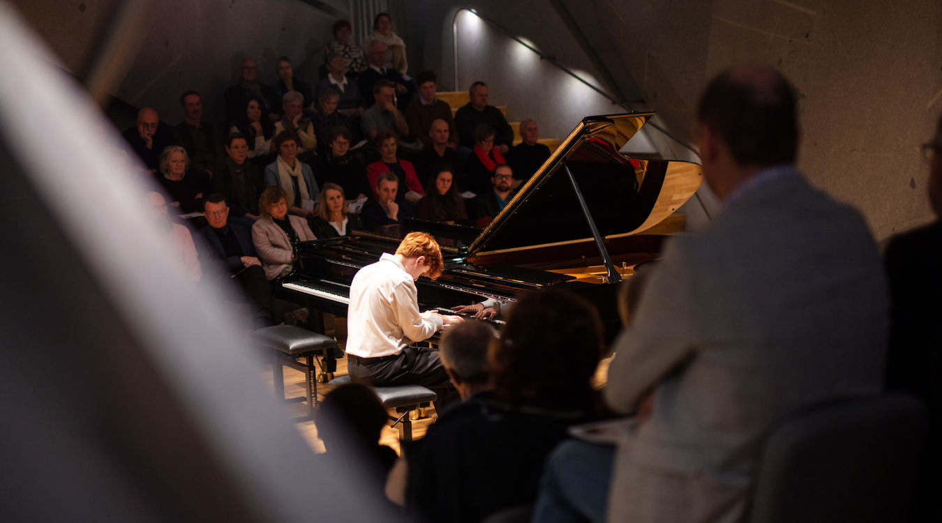 Klavierkonzert Haus Marteau Ein junger Mann im weißen Hemd spielt auf einem Flügel vor Publikum. Der Saal ist interessant beleuchtet.