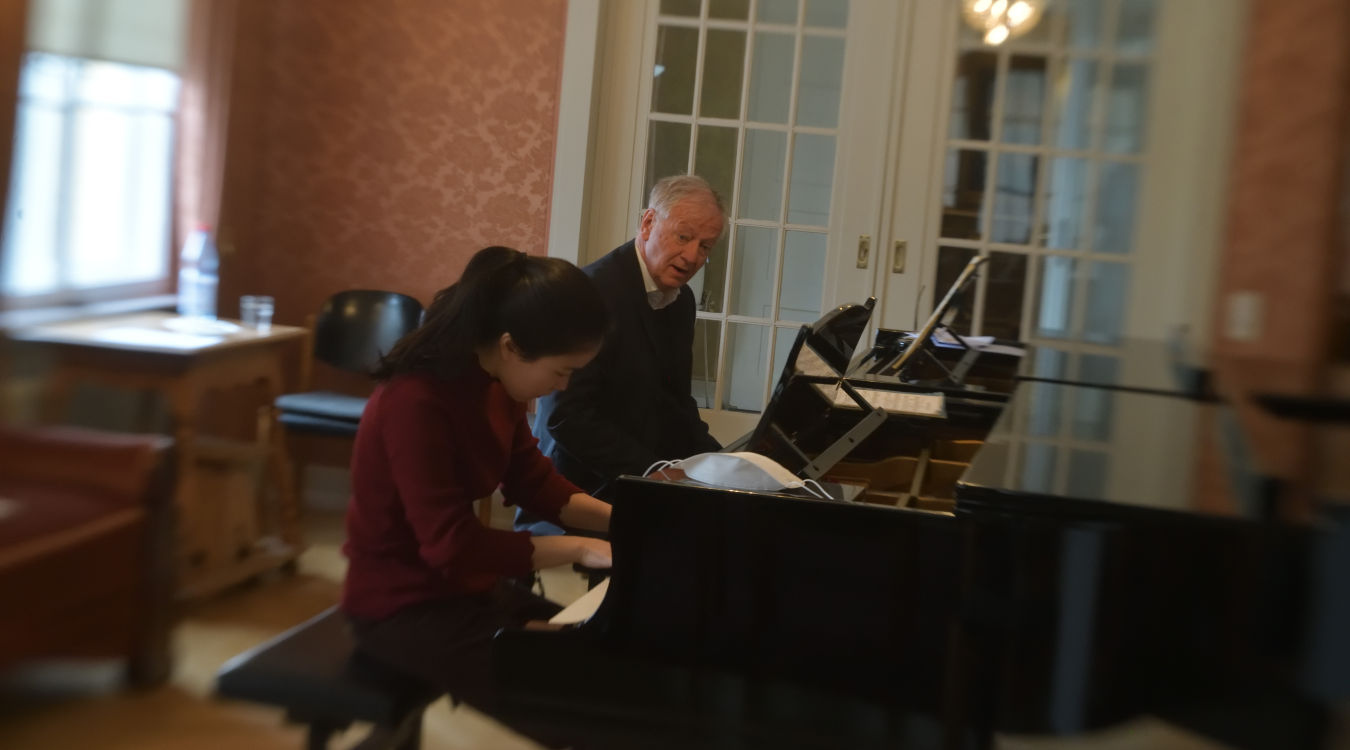 Meisterkurs für Klavier von Prof. Arnulf von Arnim In einem mit Antiquitäten ausgestattetem Raum stehen zwei Flügel, an denen ein älterer Herr und eine junge Dame zusammen spielen.