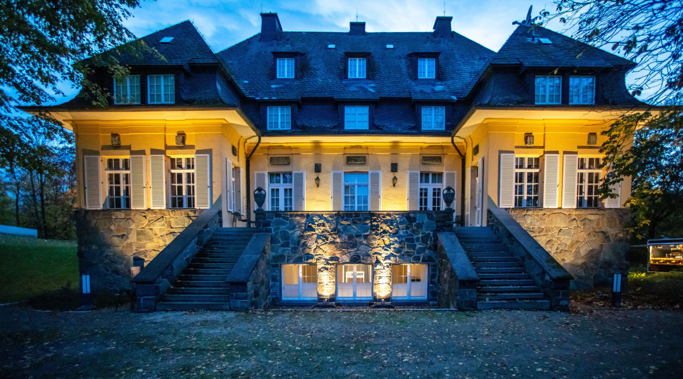 Eine alte, von Bäumen umgebene Villa ist in der Dämmerung atmosphärisch beleuchtet.