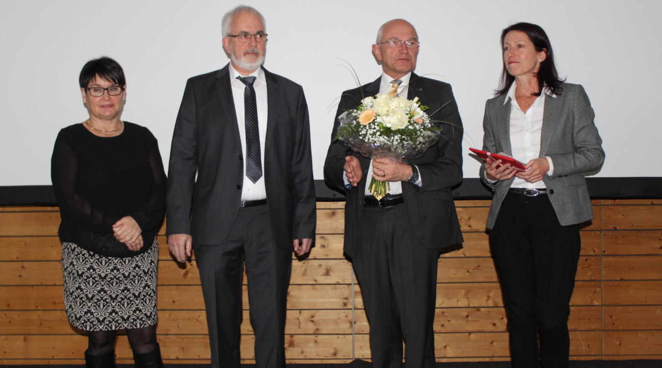 Ein bewegender Moment: Dr. Klaus Leipziger steht während seiner Verabschiedung gemeinsam mit seiner Frau auf der Bühne. Daneben sind Bezirkstagspräsident Dr. Günther Denzler mit einem Blumenstrauß und Vorstand Katja Bittner zu sehen. 