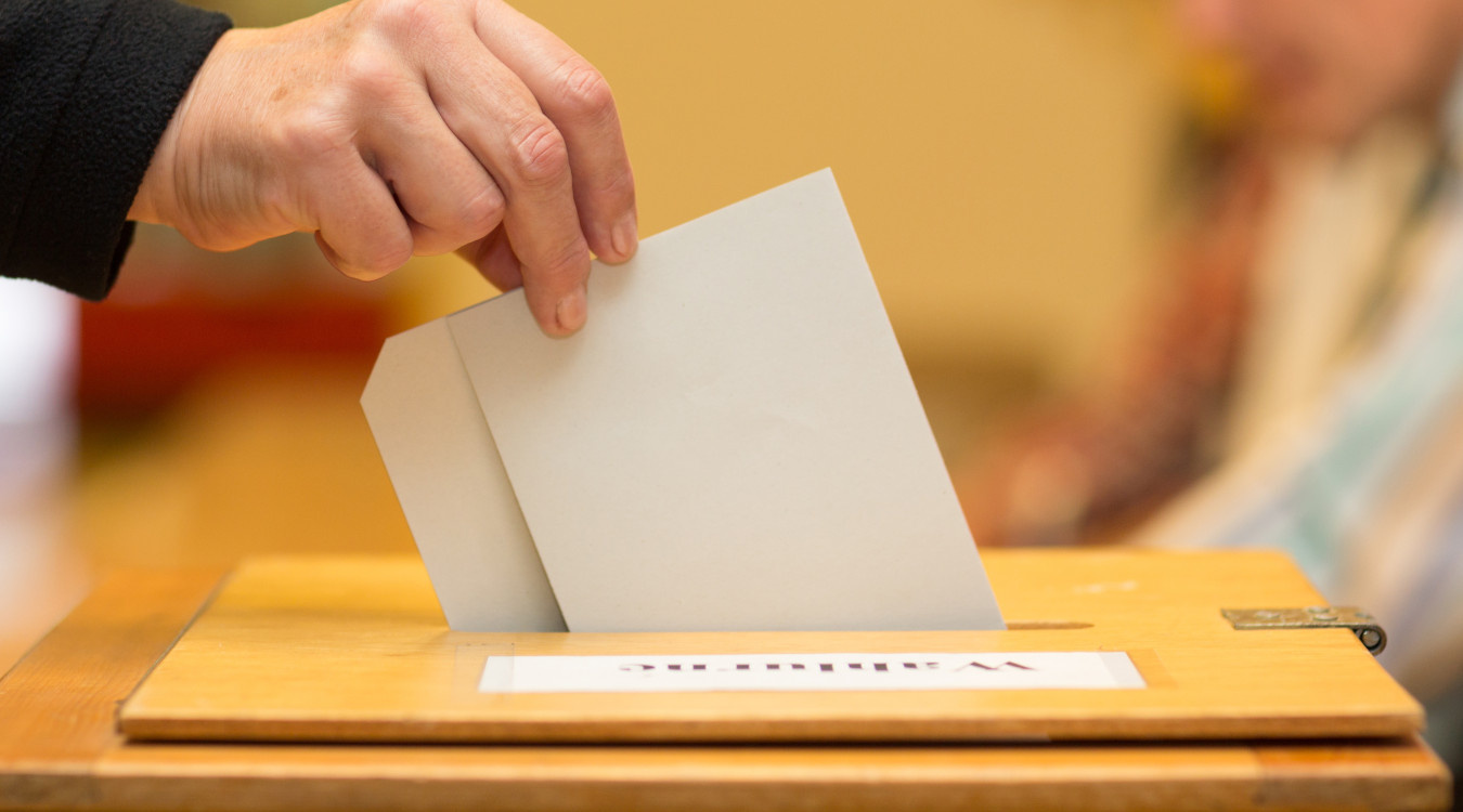Das Bild zeigt eine Wahlurne in Nahaufnahme und eine Hand, die gerade einen Wahlzettel einwirft.