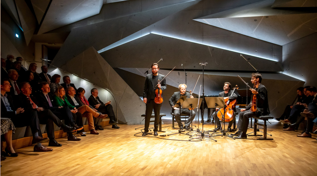 vier Musiker stehen in einem Konzertsaal, links und rechts sitzen Menschen im Publikum