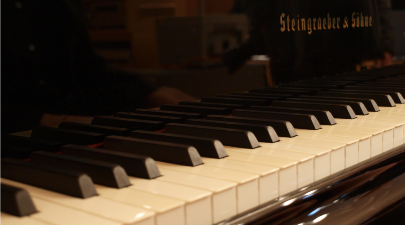 Das Bild zeigt eine Nahaufnahme eines Klaviers. Es sind die schwarz-weißen Tasten zu sehen. 