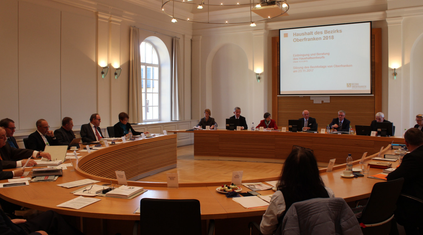 Der Bezirkstag von Oberfranken tagt in einem hellen Sitzungssaal. Das Bild zeigt die Bezirksräte auf ihren Plätzen, im Mittelpunkt ist Bezirkstagspräsident Dr. Günther Denzler zu sehen.