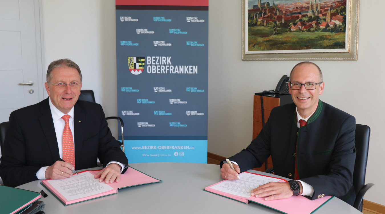 Links sitzt Bezirkstagspräsident Henry Schramm und rechts Landrat von Lichtenfels Christian Meißner. Beide halten einen Stift in der Hand und unterschreiben die Kooperationsvereinbarung.