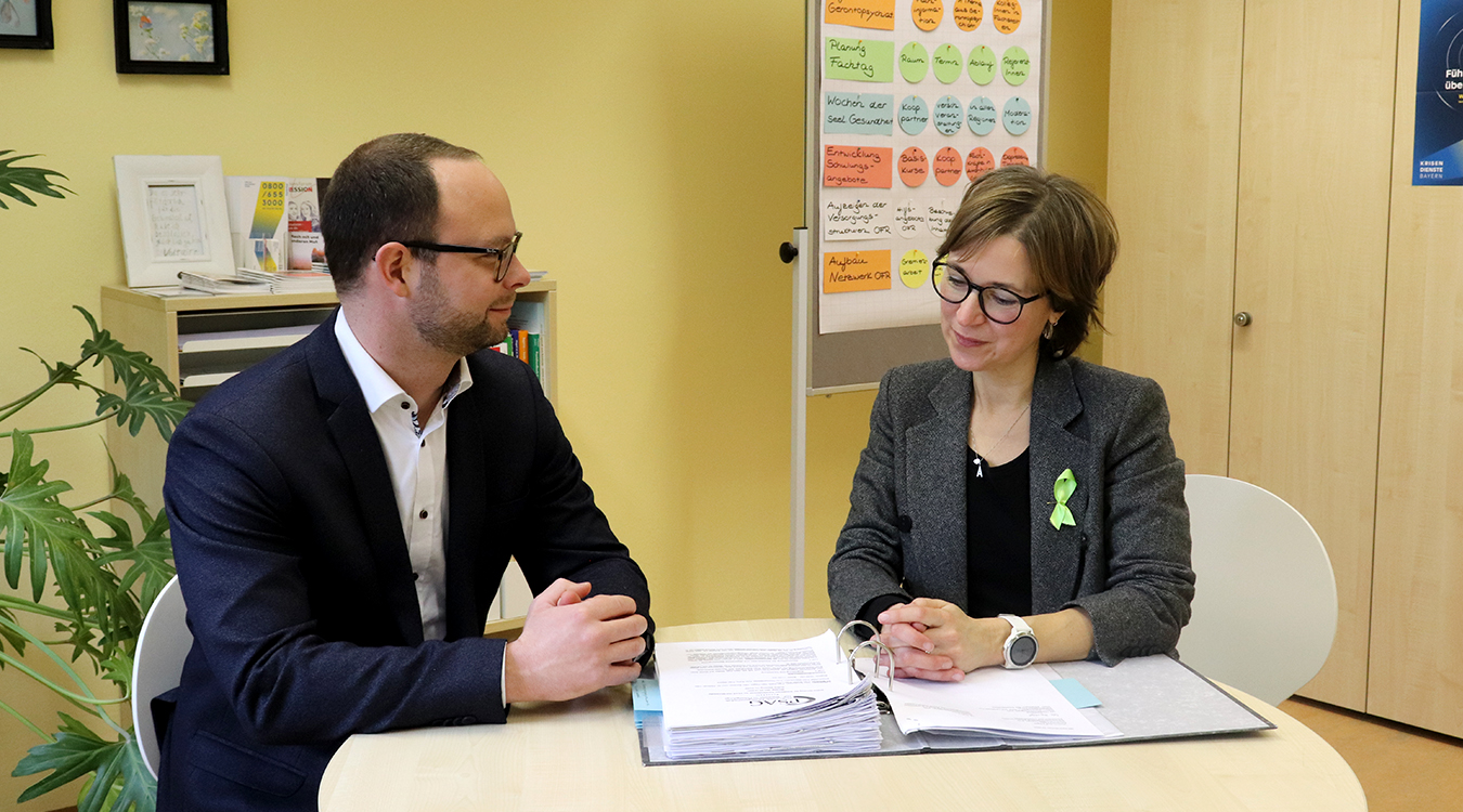 Alexandra Pape und Norbert Neumüller Eine Frau und ein Mann in Anzug sitzen nebeneinander an einem Tisch, auf dem Unterlagen liegen.