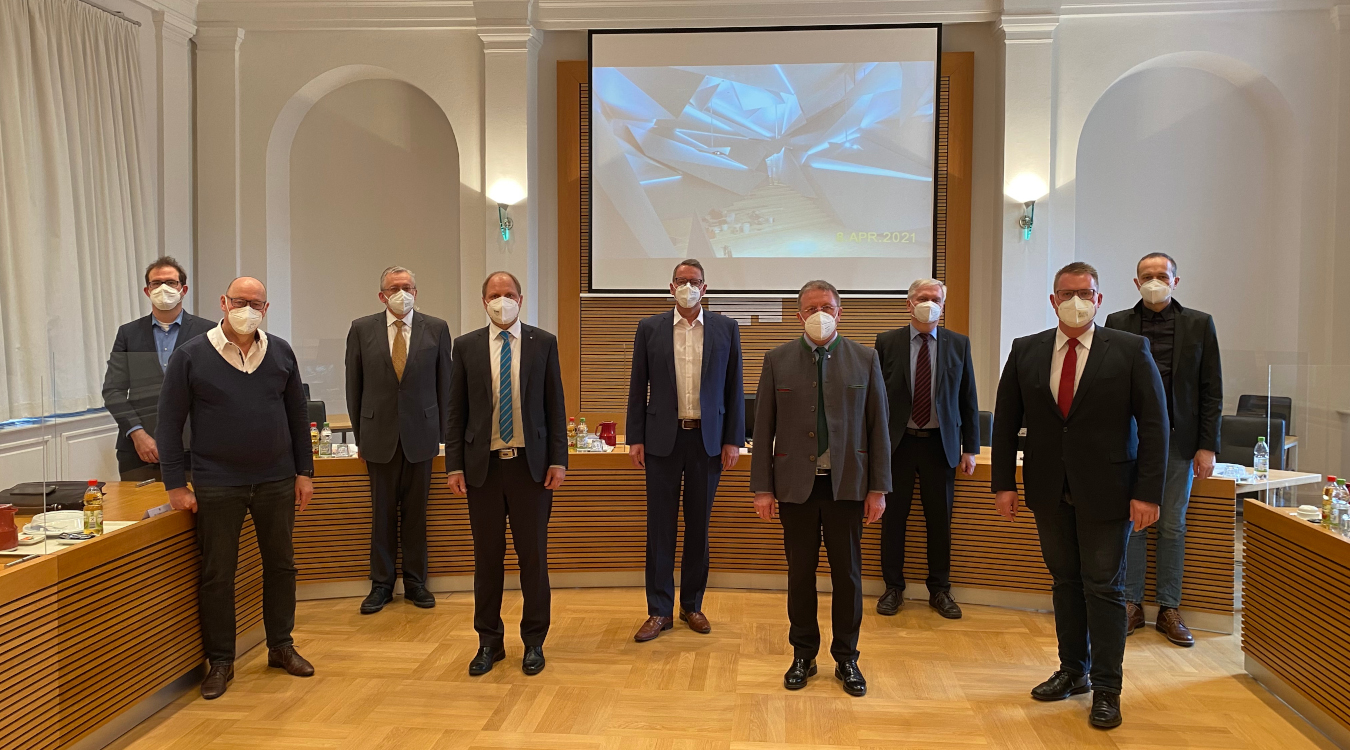 Neun Männer in Anzug und mit Mund-Nasen-Bedeckung stehen in einem Sitzungssaal.