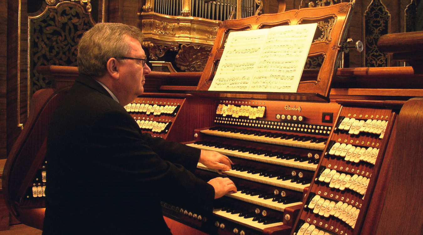 Ein älterer Mann spielt auf einer Orgel. Im Hintergrund prunkvolle Goldverzierungen an den Wänden zu erkennen.