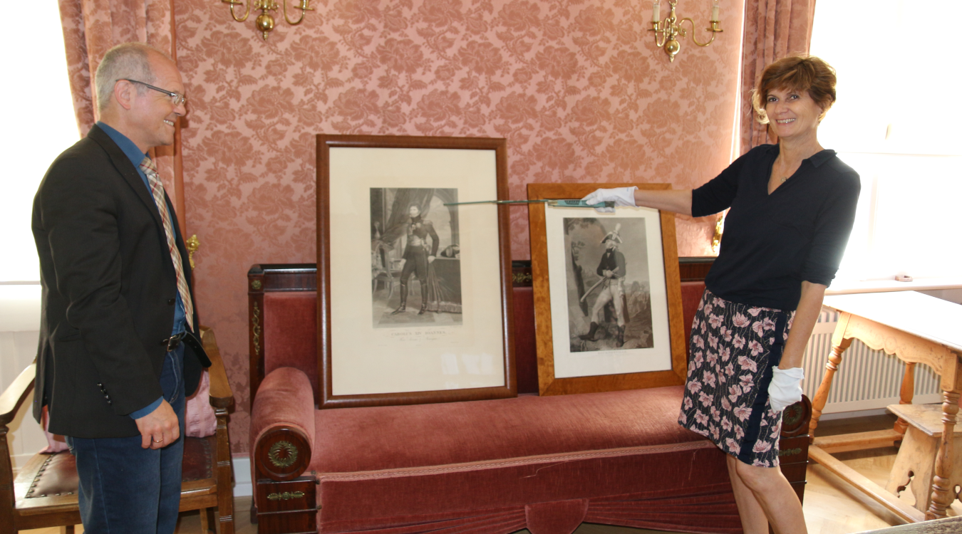Verwaltungsleiter Dr. Ulrich Wirz und Restauratorin Sabine Schumm sind zu sehen sowie zwei Bilder, die auf einer historischen Couch stehen.