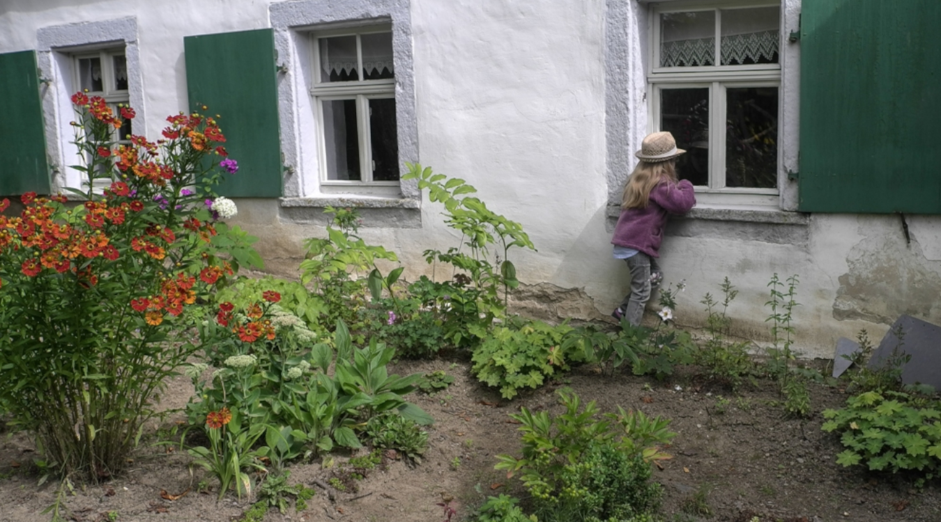 Maus-Türöffner-Tag Ein Mädchen steht in einem Garten und schaut durch ein Fenster in ein altes Haus hinein.
