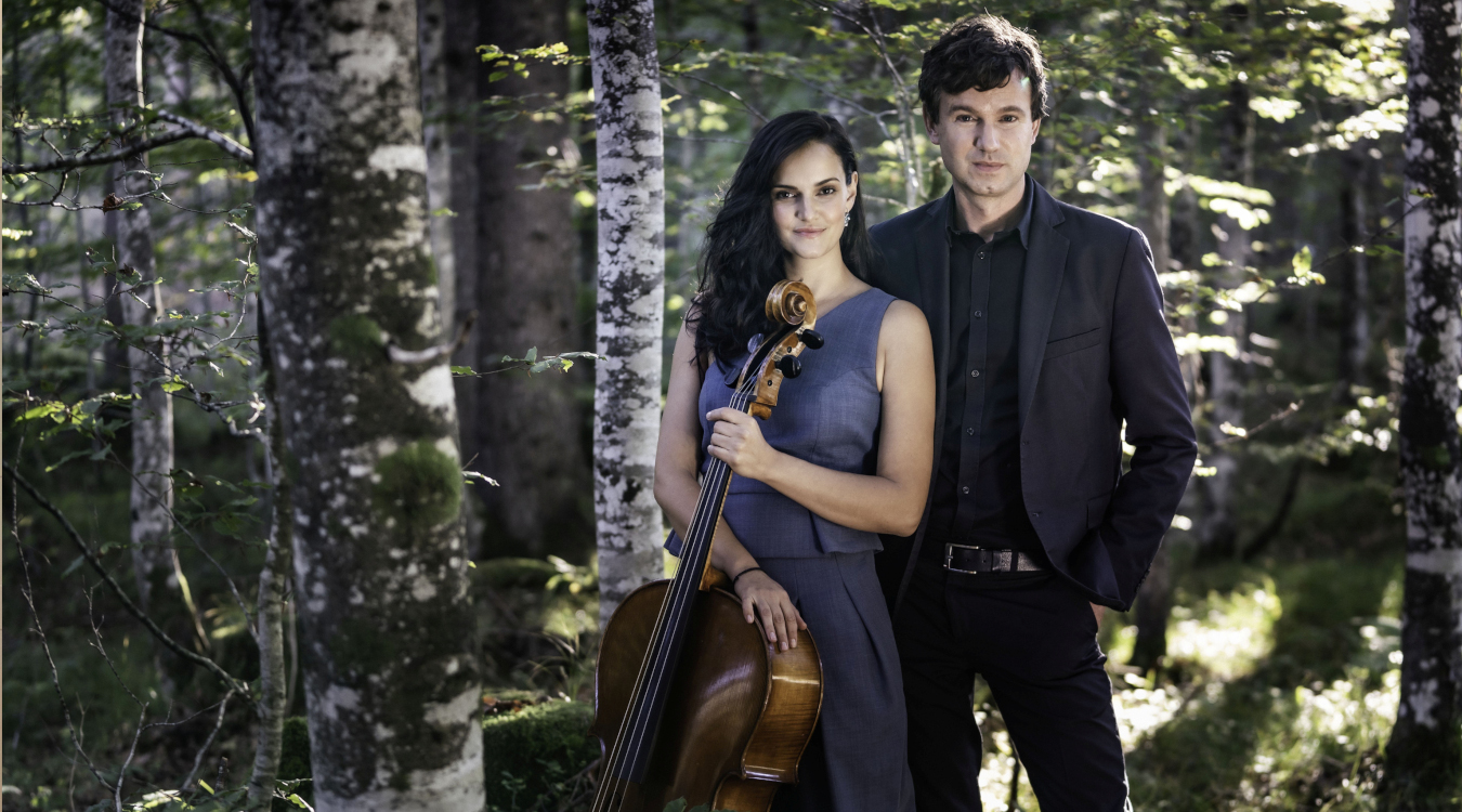 Eine Frau im Abendkleid mit Cello steht in einem Wald, hinter ihr steht ein Mann in einem Anzug