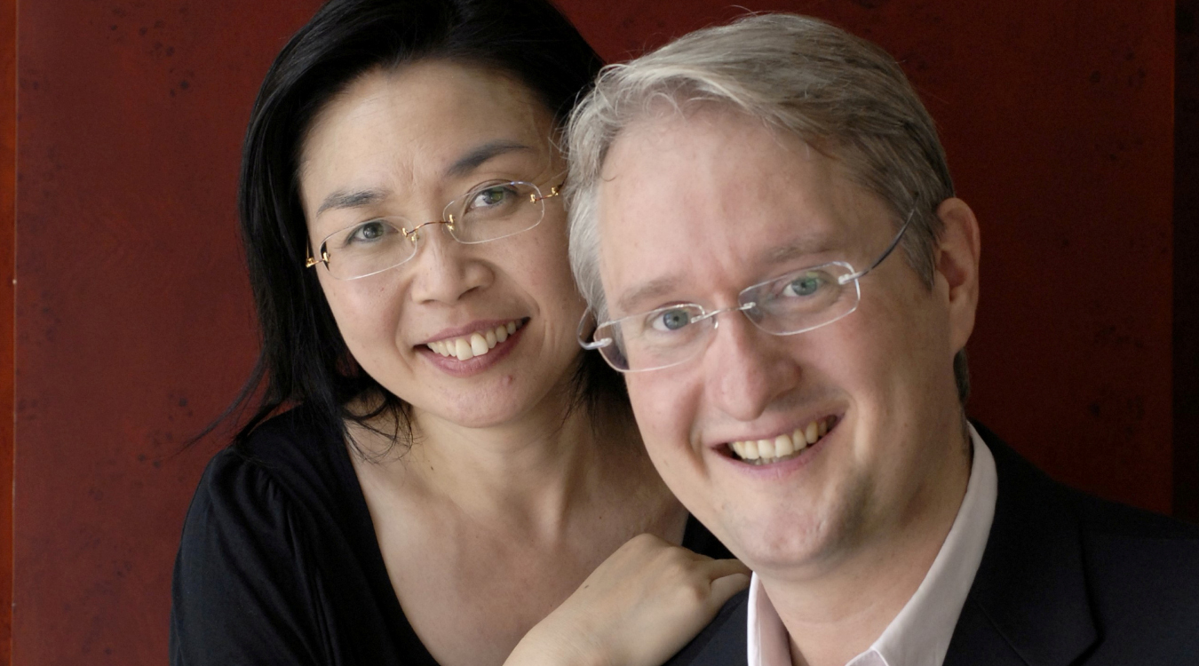 Eine Frau mit Brille lehnt sich auf der Schulter eines Mannes mit Brille und weißem Haar auf. Beide lächeln. 