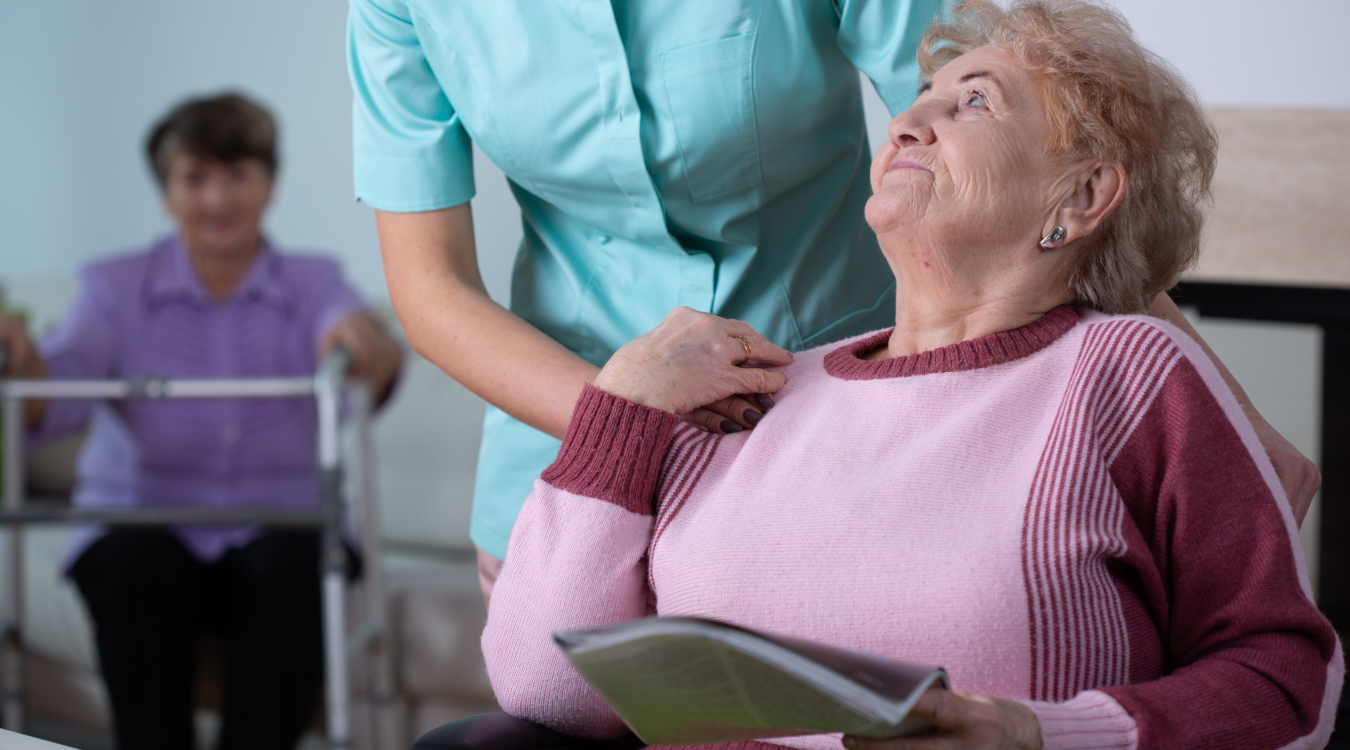 Eine Seniorin hält eine Zeitschrift in der Hand. Hinter ihr steht eine Pflegerin und legt eine Hand auf ihre Schulter.