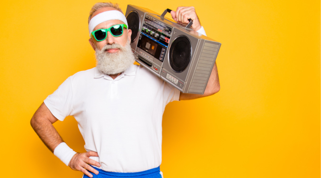 Das Bild zeigt einen Senior mit weißem Rauschebart, Sonnenbrille, weißem T-Shirt und einer kurzen Hose in blau vor einem goldgelben Hintergrund. Er trägt lässig ein altes Kassetten-Radio über der linken Schulter.